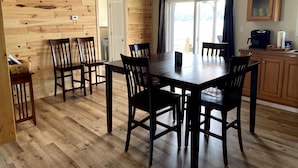 A dining area with a wooden table and chairs, a kitchen area with cabinets and a coffee machine, and a view of the outdoors through a window. - Sunrise, Sunset, Relax, Repeat Lamoka Cove (Bradford)