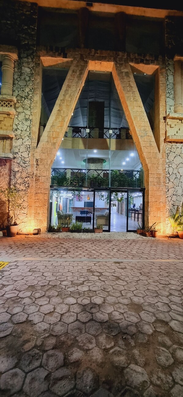 Entrada de un edificio con arco de piedra, plantas en macetas y un camino empedrado que conduce a un espacio interior con mesas y sillas.