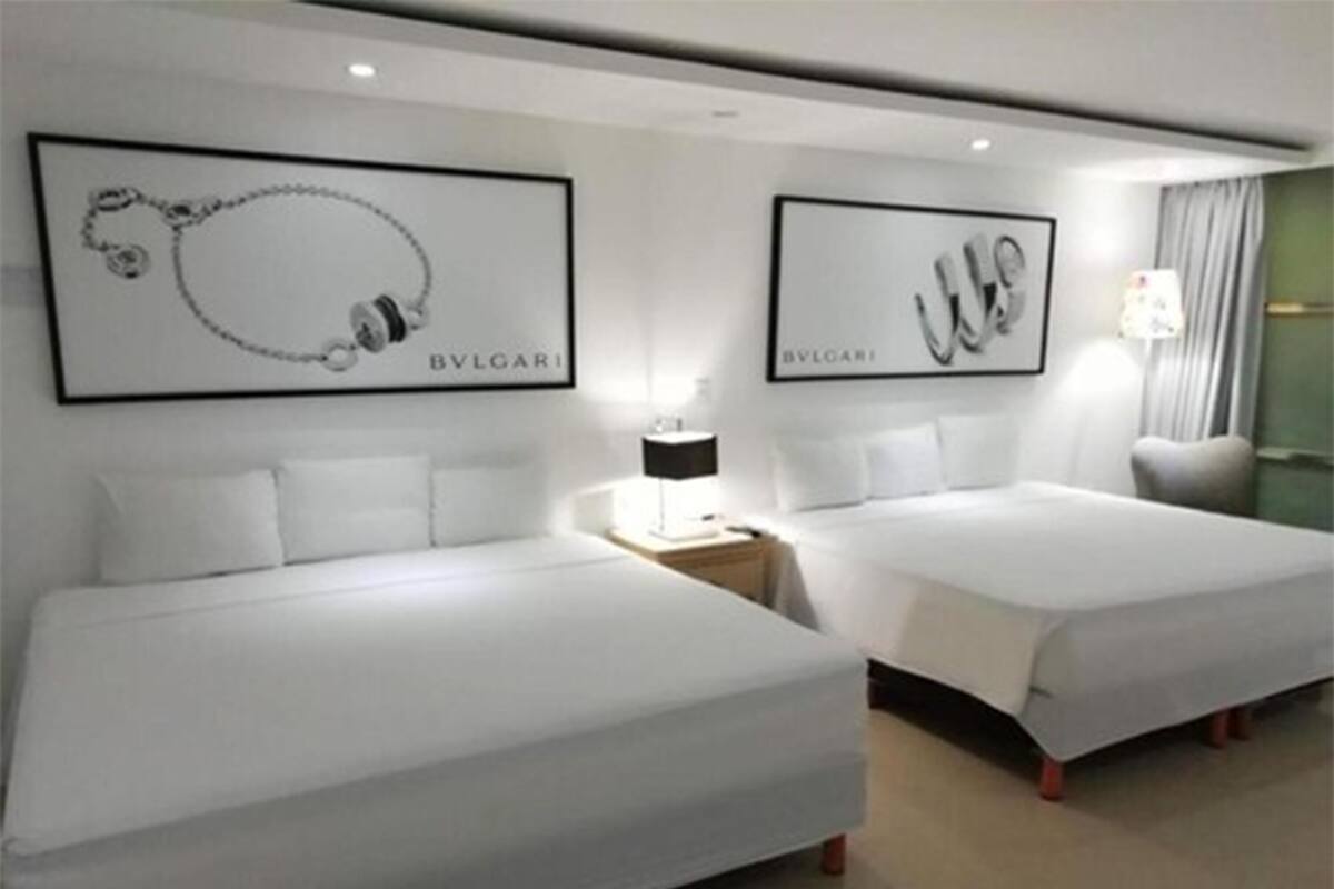 A hotel room with two beds, a nightstand with a lamp, and two framed pictures of a necklace and a watch on the wall.