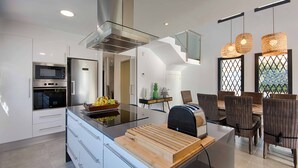 A modern kitchen with a central island, built-in oven, and refrigerator. There is a dining area with wicker chairs and a table. - TER MARAVILLOSA VILLA CANAL TER,PISCINA,WIFI, PARKING (Empuriabrava)