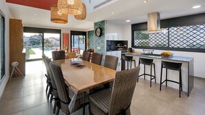 A modern kitchen with a dining area, featuring a large wooden table and wicker chairs, a built-in stove, and a view through large windows. - TER MARAVILLOSA VILLA CANAL TER,PISCINA,WIFI, PARKING (Empuriabrava)
