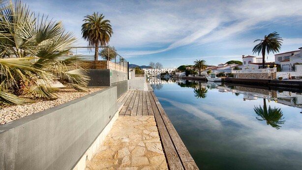 Une promenade en bord de mer, avec un sentier pavé de pierres, des palmiers et une étendue d’eau calme qui reflète le ciel.