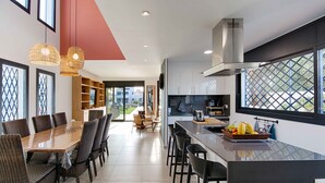 A modern kitchen with a central island, dining area with wicker chairs, and large windows offering views. - TER MARAVILLOSA VILLA CANAL TER,PISCINA,WIFI, PARKING (Empuriabrava)