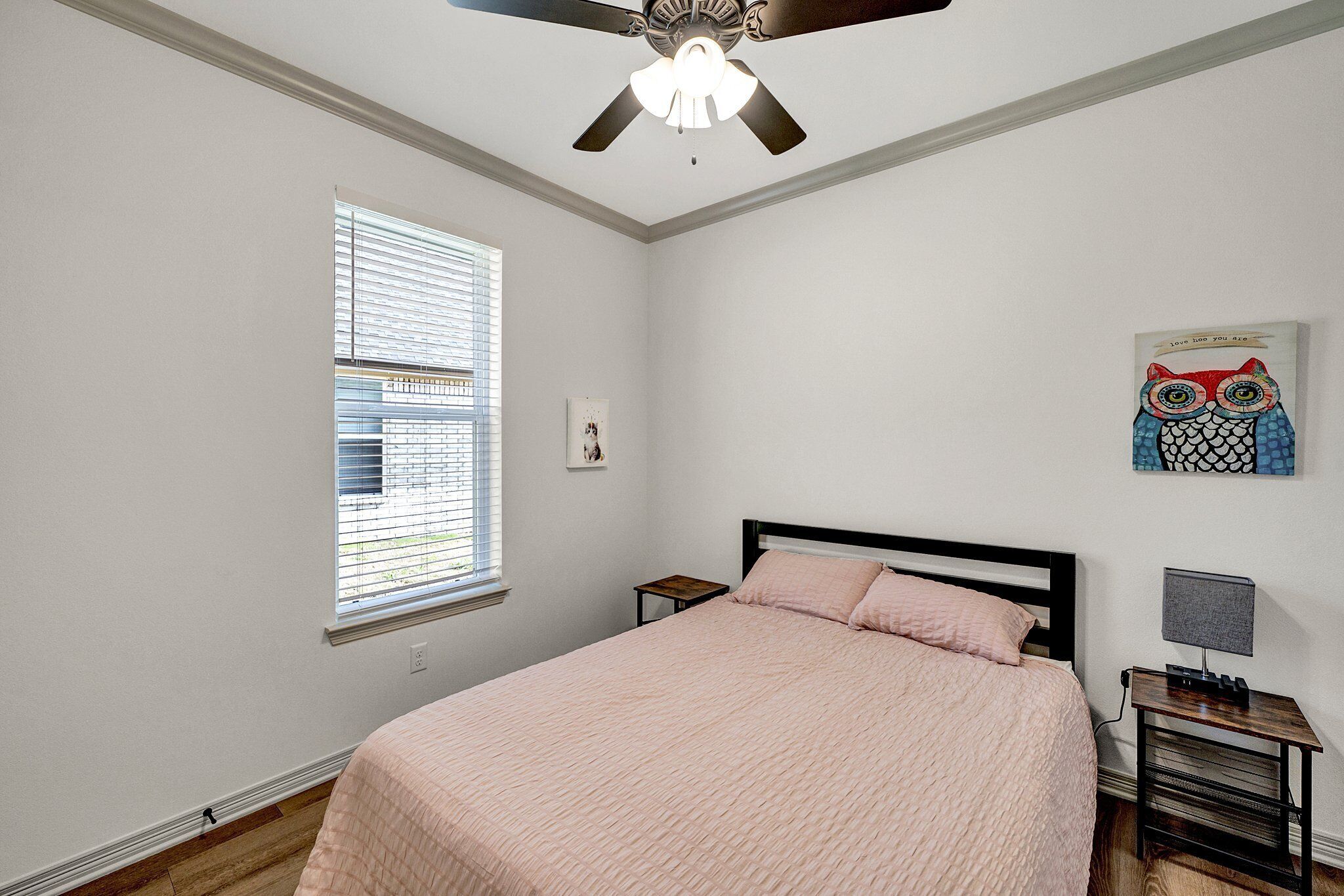 Une chambre à coucher comprenant un lit, une table de chevet, un ventilateur de plafond, une fenêtre avec des stores et une œuvre d’art accrochée au mur.