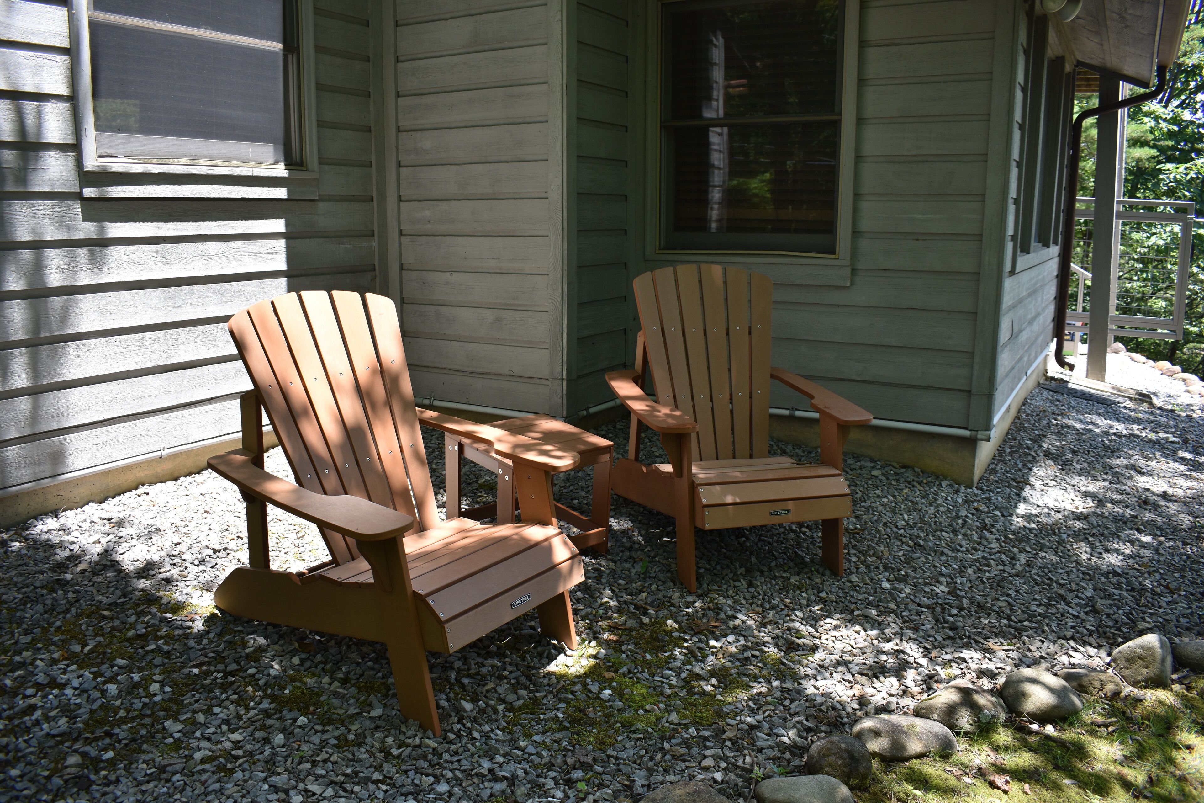 Two wooden Adirondack chairs and a small table on a gravel surface outside a house with a window.