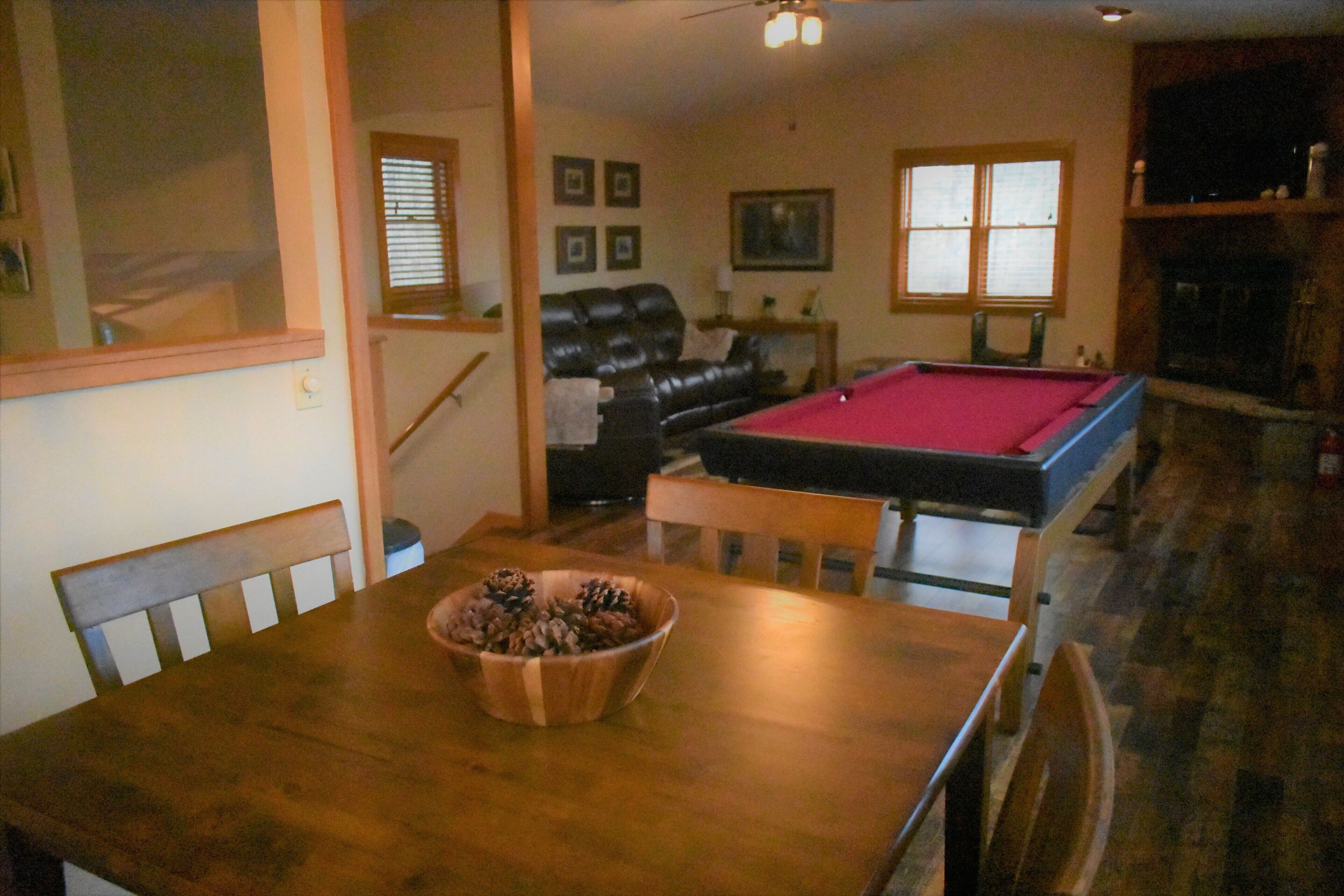 Un comedor con una mesa de madera, un cuenco de piñas y una mesa de billar al fondo.