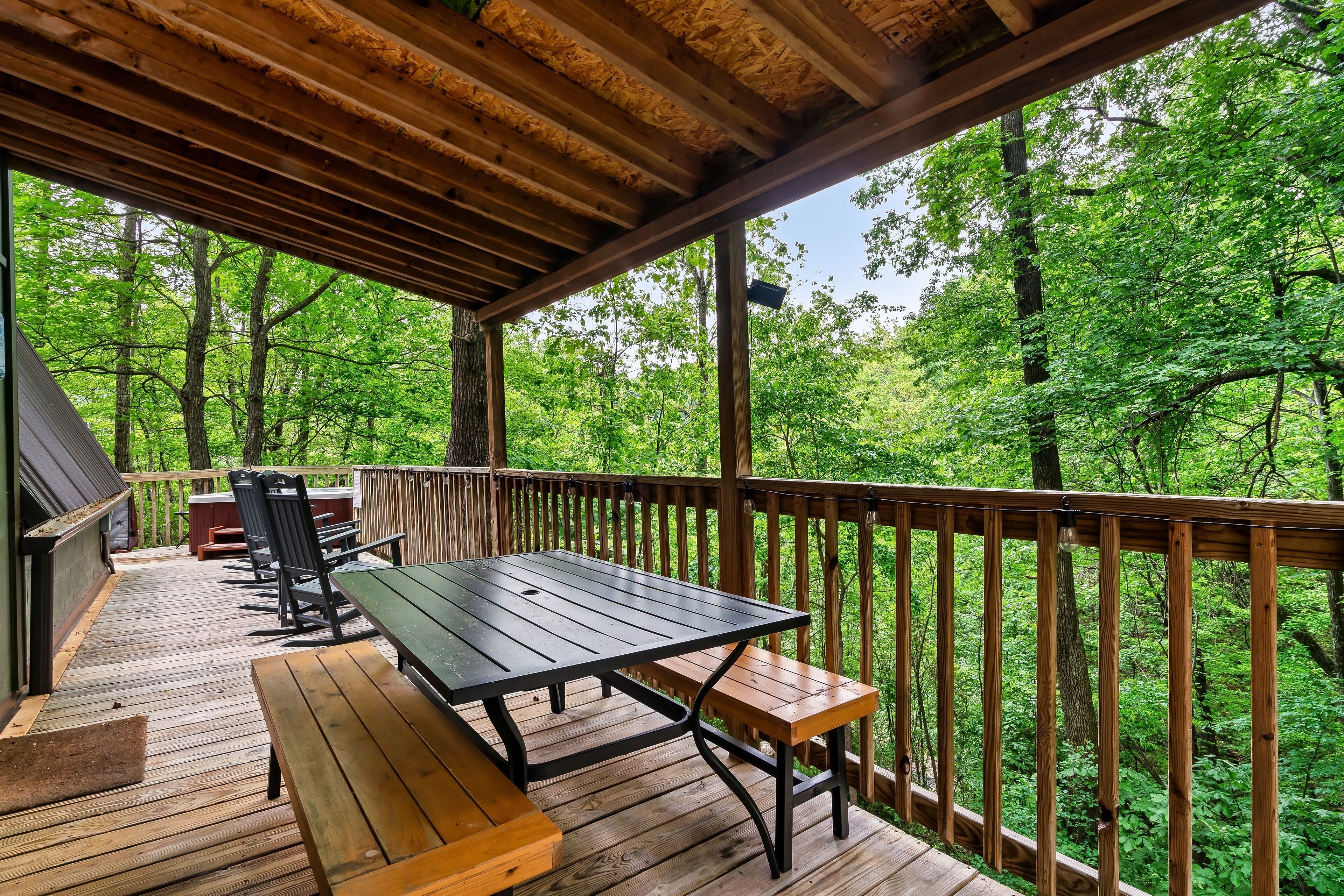 Um deck de madeira com mesa de piquenique e bancos, cercado por guarda-corpo de madeira, com vista para uma área arborizada.