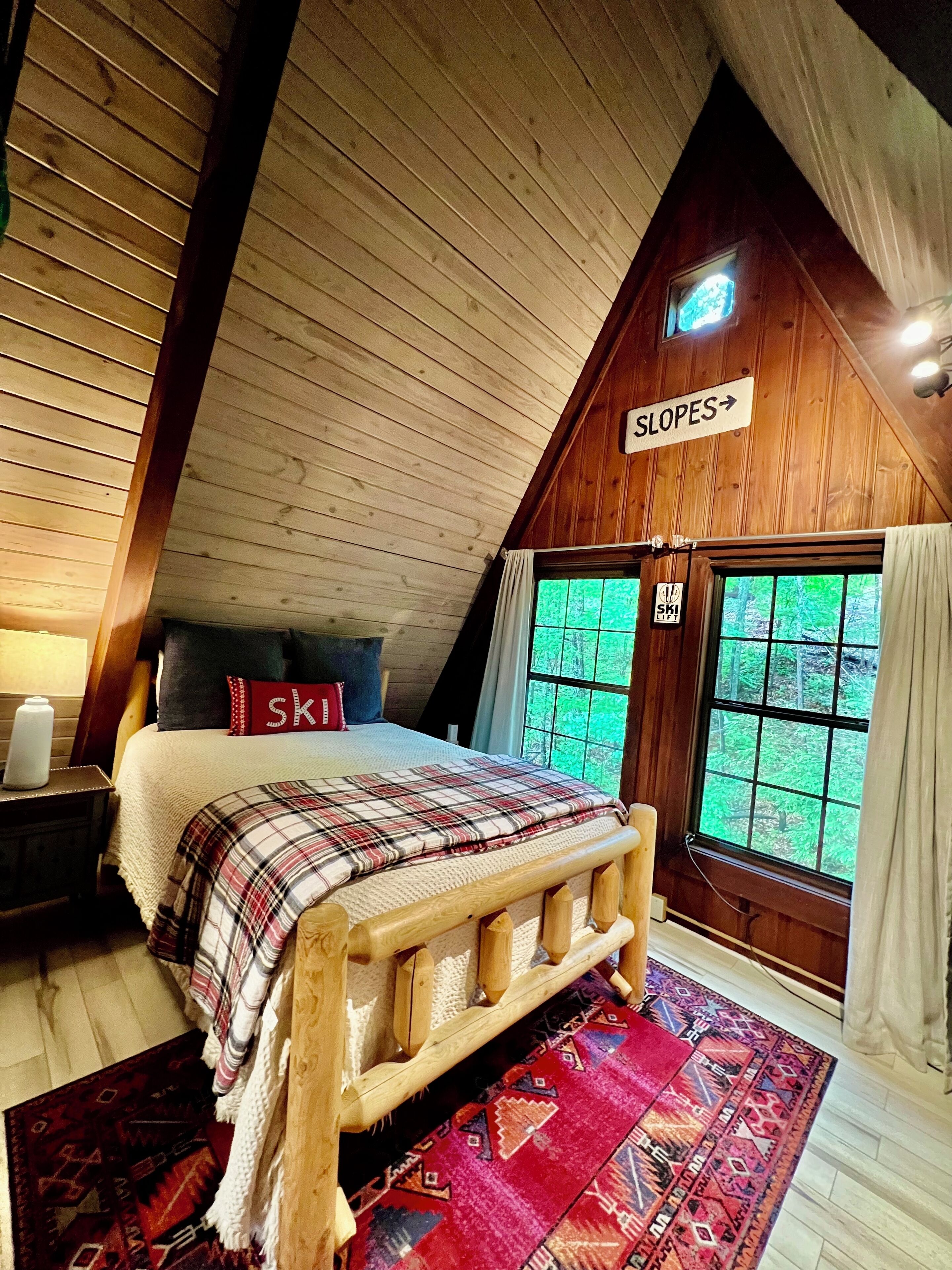 Quarto aconchegante com teto inclinado de madeira, uma cama com manta xadrez, travesseiro vermelho e janela com vista para o verde.