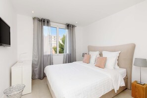 Un dormitorio con cama, televisión, escritorio, silla, lámpara y ventana con cortinas.