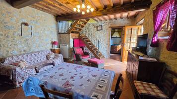 A rustic interior with a wooden ceiling, a dining table with a floral tablecloth, a sofa, and a kitchen area with a television.