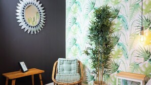 Una habitación con pared oscura, un papel tapiz con hojas grandes, techo con estructura de madera, una silla de mimbre, un tapete redondo y una mesita con un marco de fotos.