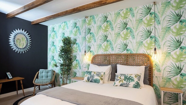 Un dormitorio con cama, cabecera de ratán, pared con papel tapiz de hojas tropicales, espejo redondo y viga de techo de madera.