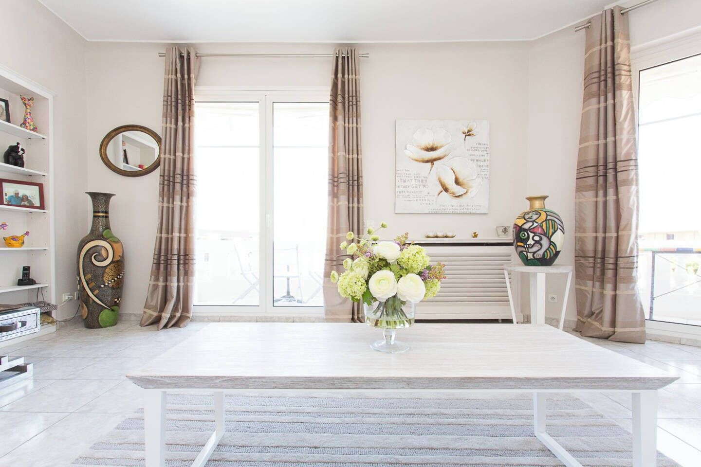 A modern living room with a white table, a vase of flowers, a fireplace, and a large window with curtains.