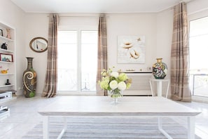 A modern living room with a white table, a vase of flowers, a fireplace, and a large window with curtains. - Croisette Boulevard deluxe apartment (Cannes)