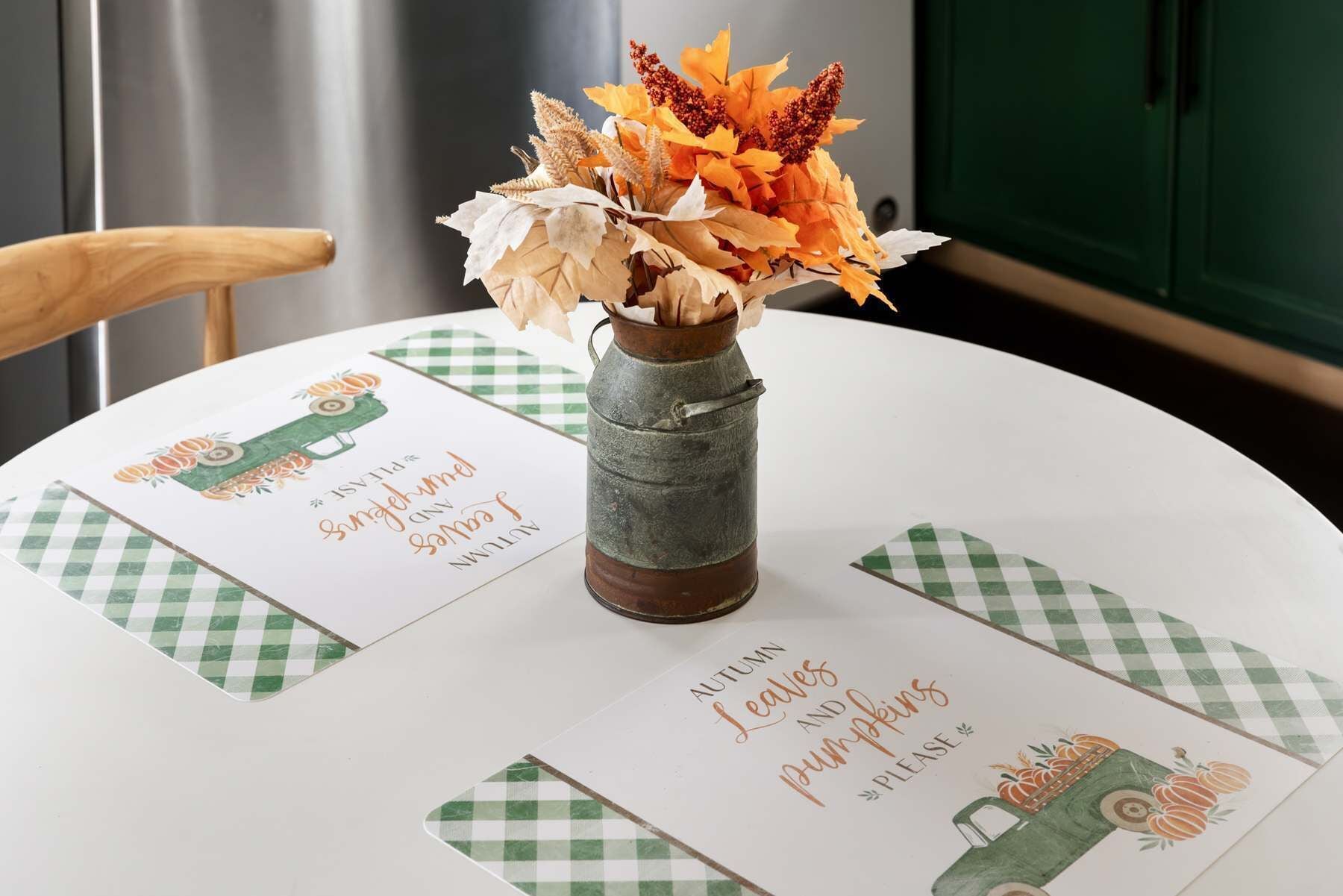 A table with a checkered tablecloth, a rustic metal vase with autumn leaves, and a placemat with autumn-themed illustrations and text.
