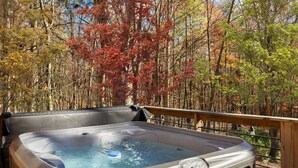 A hot tub on an outdoor deck with a view of autumn trees. - Cozy Cabin w Lake, Hot Tub, Huge Deck & Fire Pit (Mathias)
