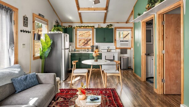 A compact living space with a kitchenette, a dining area, and a seating area with a sofa and chairs. - Cozy Cabin w Lake, Hot Tub, Huge Deck & Fire Pit (Mathias)