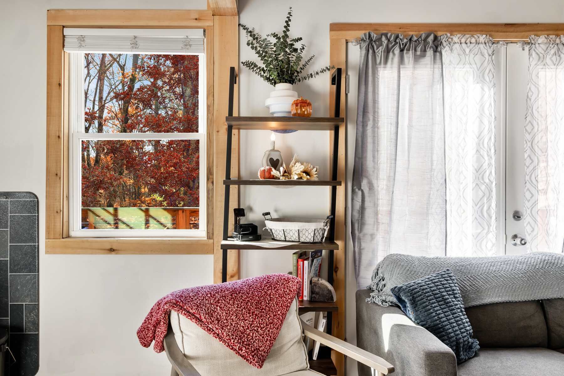 A cozy living room with a fireplace, a bookshelf, and a view of autumn trees through a window.