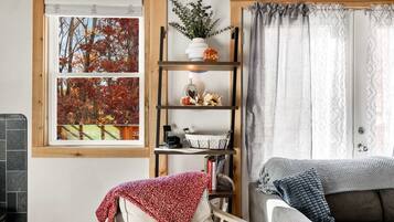 A cozy living room with a fireplace, a bookshelf, and a view of autumn trees through a window.