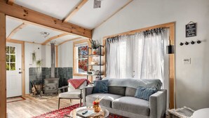 A cozy living room with a grey sofa, a round glass coffee table, and a red patterned rug. - Cozy Cabin w Lake, Hot Tub, Huge Deck & Fire Pit (Mathias)