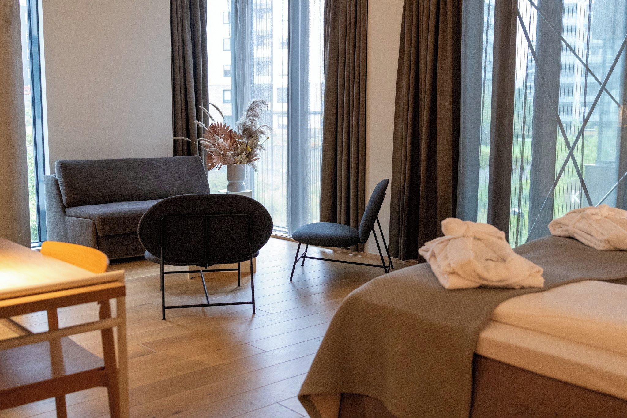 Hotellihuone, jossa on sohva, tuoli, pyyhkeillä varustettu sänky, pieni pöytä ja kuivakukkia sisältävä maljakko.