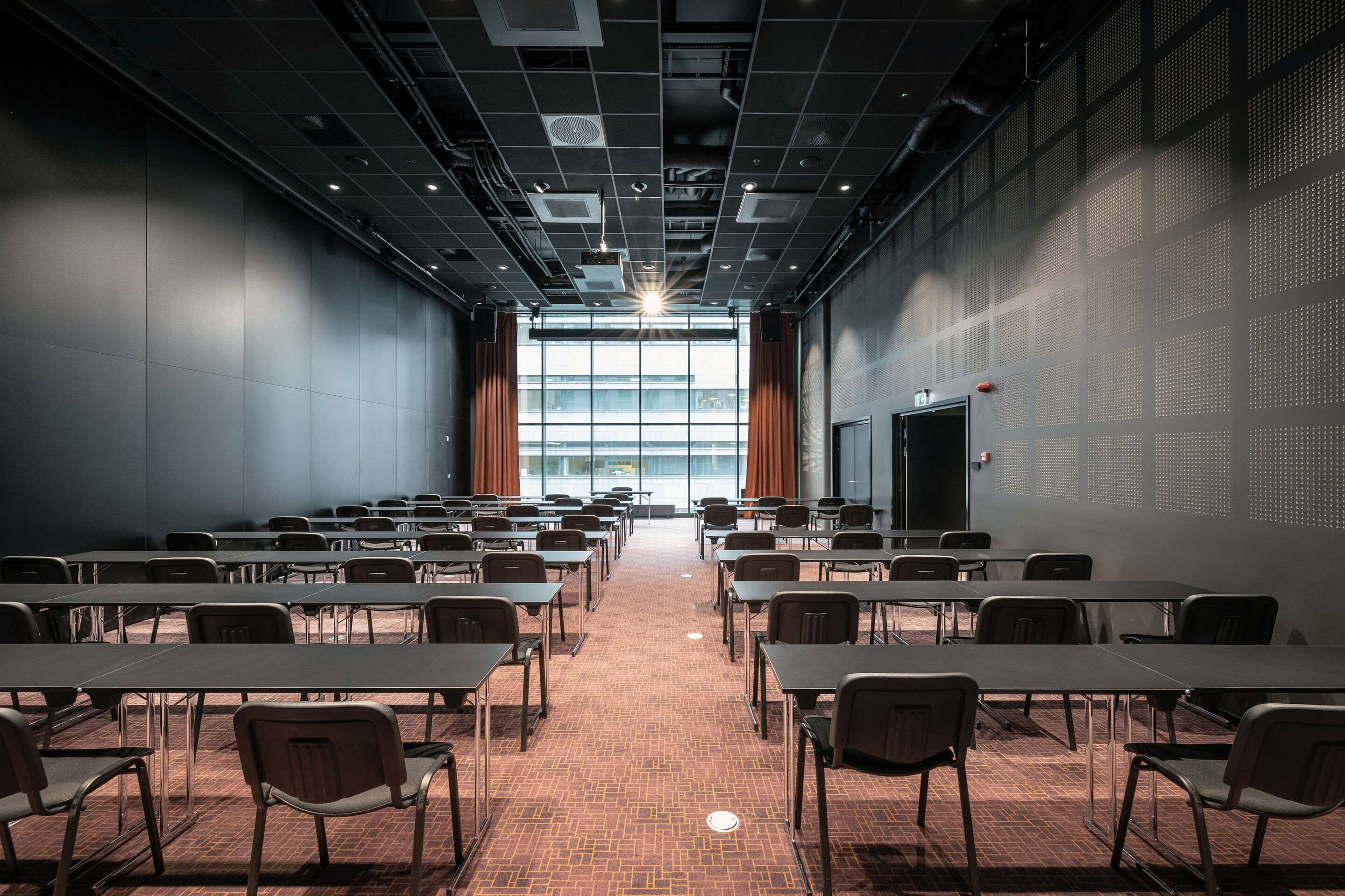 Un salón de conferencias con filas de mesas y sillas, piso de ladrillo y un ventanal con cortinas.