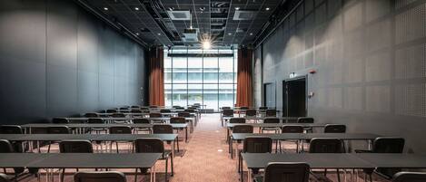 A conference room with rows of tables and chairs, a brick floor, and a large window with curtains.