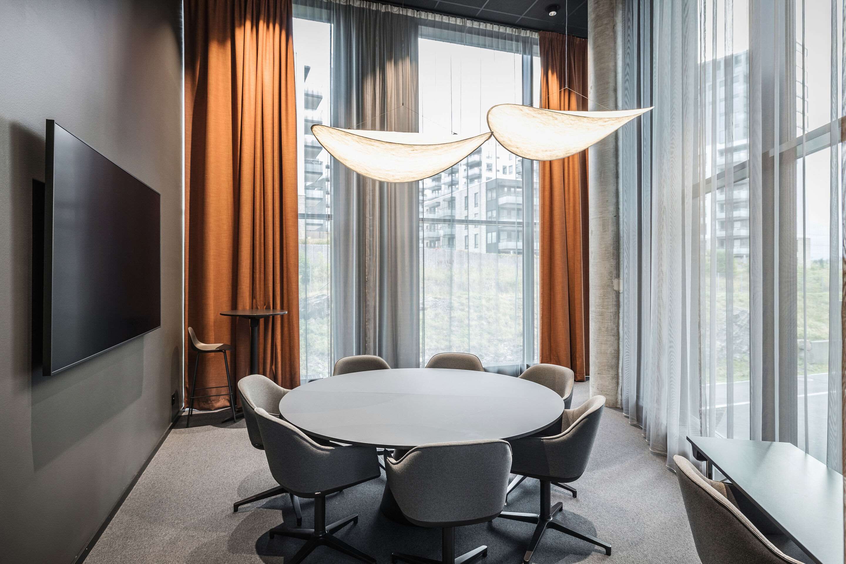A conference room with a round table, six chairs, a TV on the wall, and large windows with curtains.