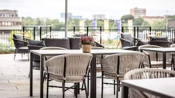 Un espace extérieur aménagé avec des chaises et des tables en osier, un panier tressé rempli de fleurs et une vue sur des bâtiments situés de l’autre côté d’une étendue d’eau.