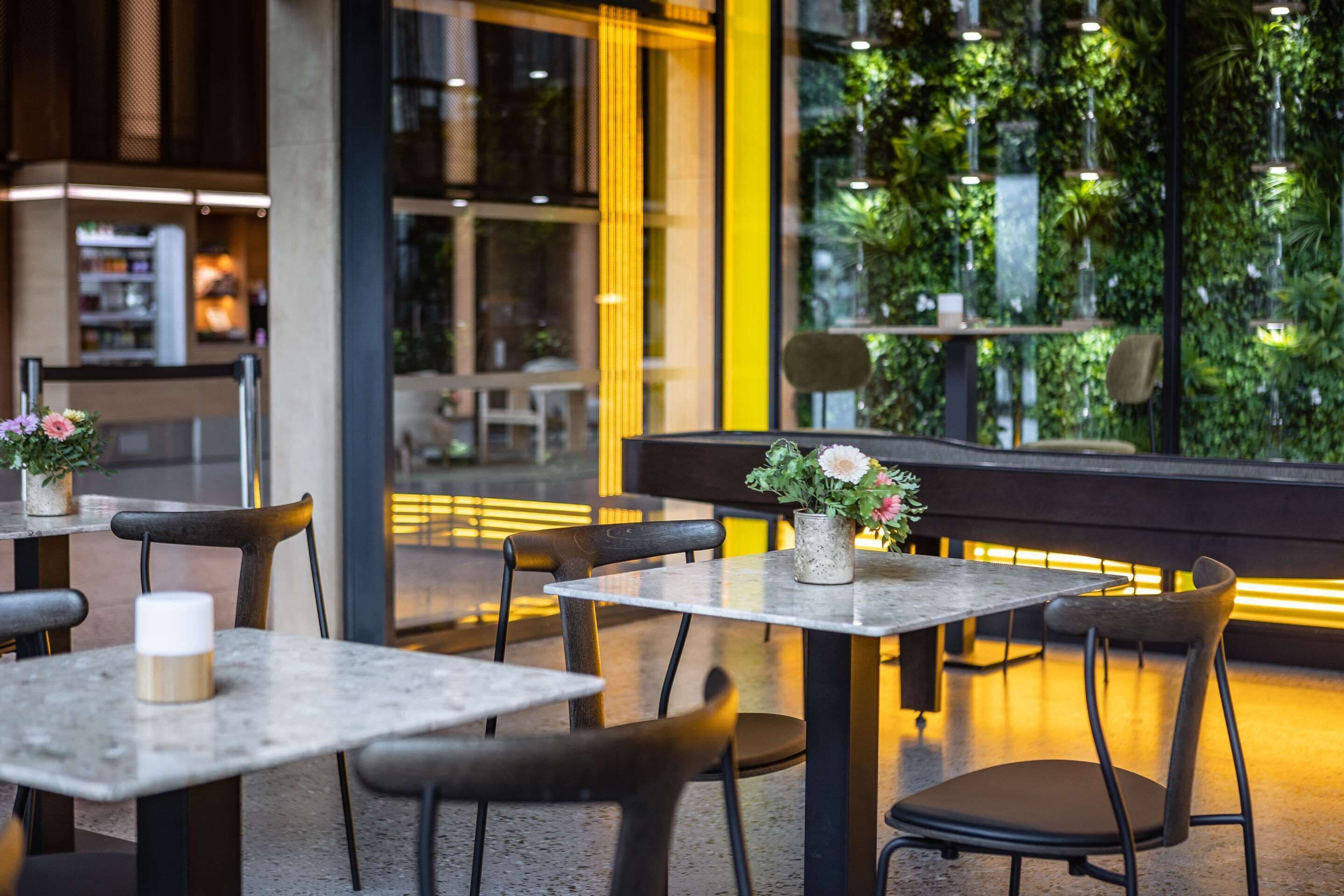 An indoor dining area with tables and chairs, a bench, and a view of indoor greenery through large windows.