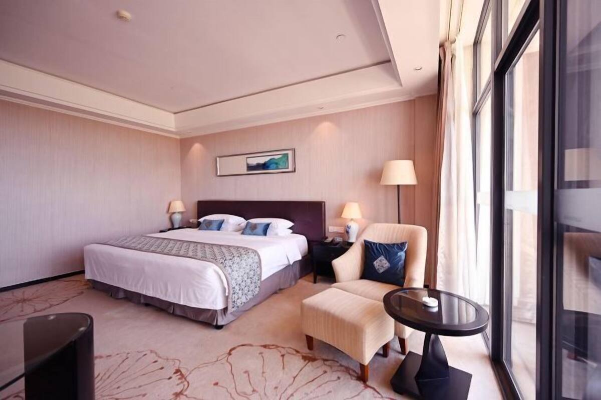 A hotel room with a large bed, two bedside tables with lamps, a chair, a small table, and a large window offering a view.