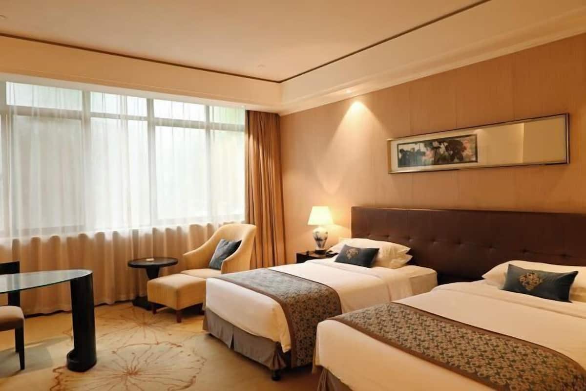 A hotel room with two beds, a sitting area with a table and chairs, a large window with curtains, and a framed picture on the wall.