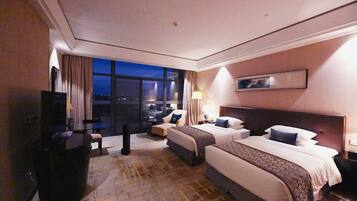 A hotel room with two beds, a desk, a TV, and a view of the city at night.