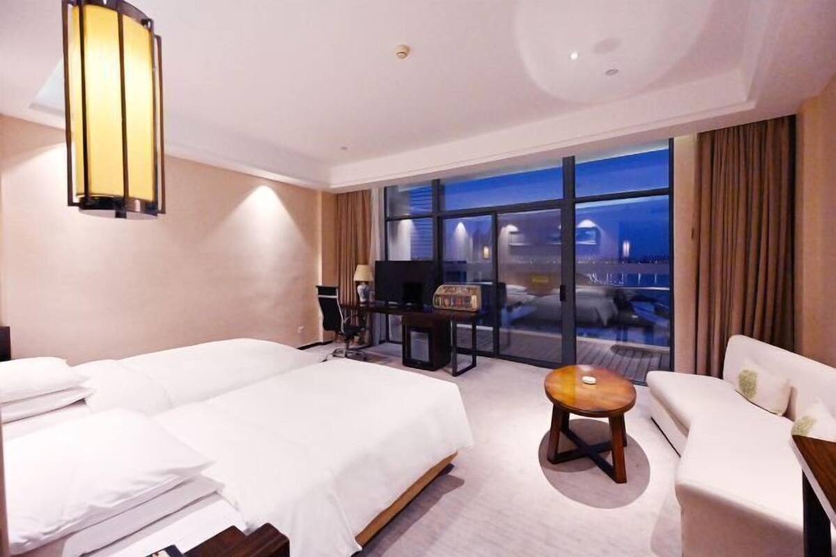 A hotel room with two beds, a sofa, a small table, a TV, and a large window with a view of the city at night.
