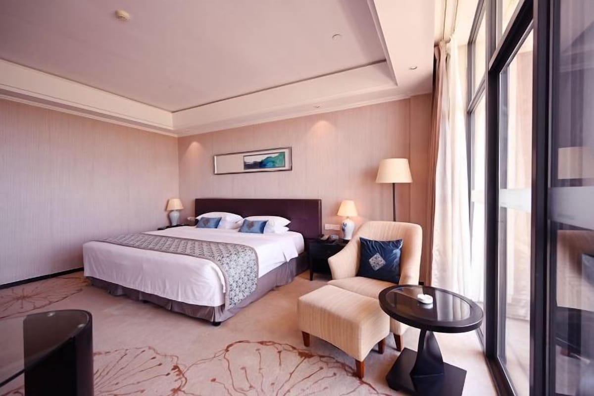 A hotel room with a large bed, two bedside tables with lamps, a chair, a small table, and a large window offering a view.
