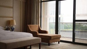 A hotel room with a large bed, a small armchair, a bedside table with a lamp, and a view of a building through a large window. - Jiuhua Chunshenhu Suzhou (Suzhou)