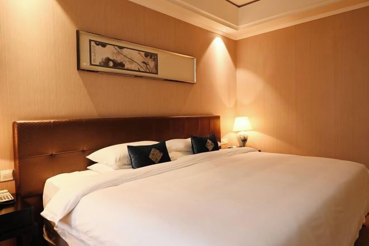 A hotel room with a large bed, a headboard, a bedside table with a lamp, and a framed picture on the wall.