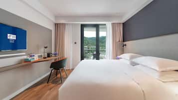 A hotel room with a bed, a desk with a chair, a television, and a view of a mountainous landscape.