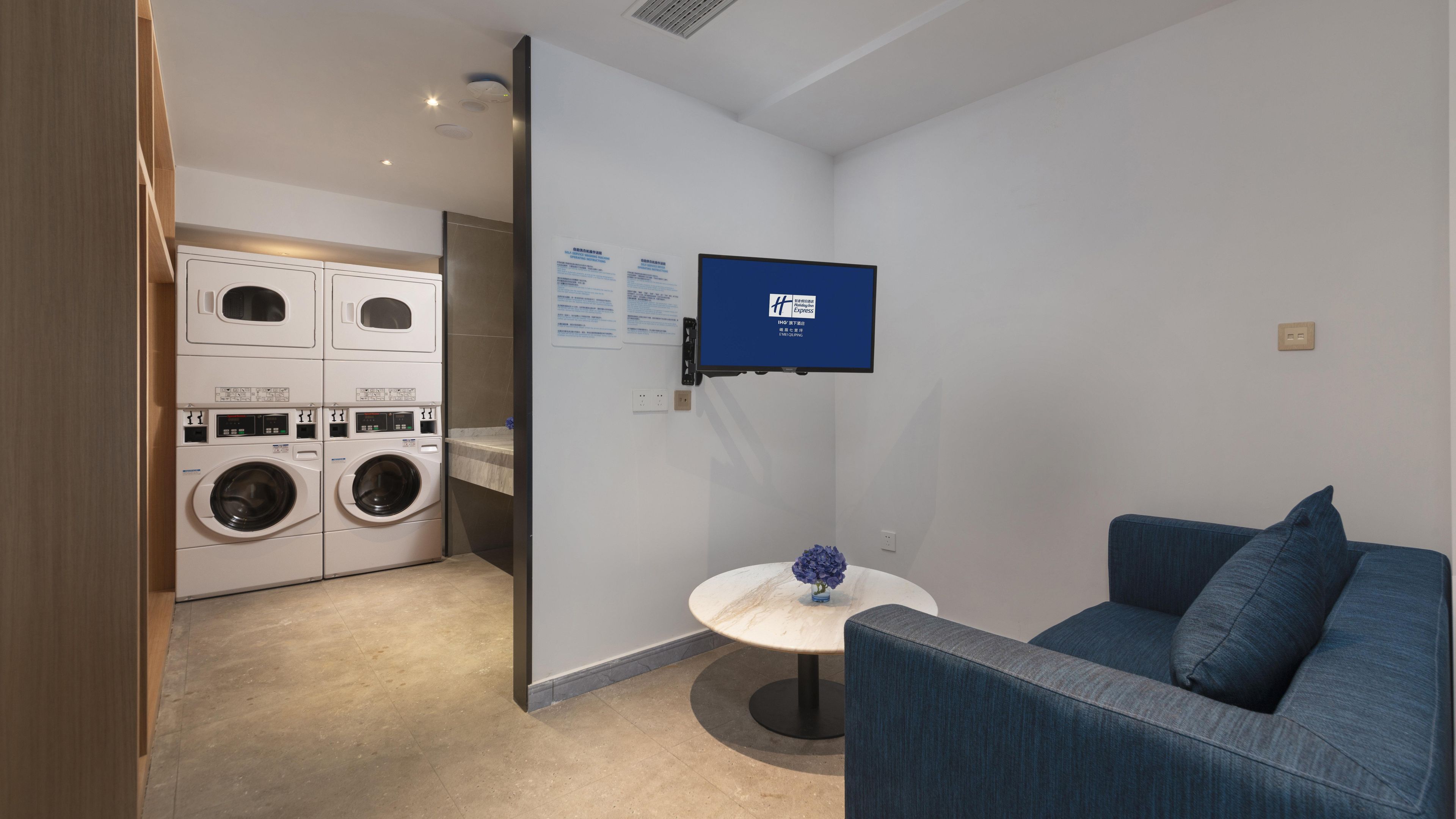 Habitación de hotel compacta con lavadora, una pequeña mesa redonda, un sofá azul y un televisor montado.