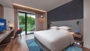 A hotel room with a bed, a desk, a chair, a small table, a nightstand, a vase with flowers, and a balcony with a view of trees. - Holiday Inn Express E'mei Qiliping (Leshan)