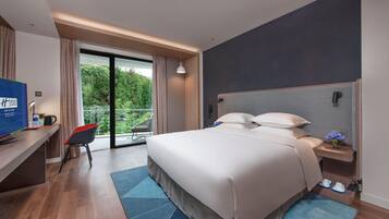 A hotel room with a bed, a desk with a computer, a chair, a nightstand, and a view of greenery outside.