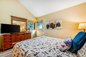 A bedroom with a bed, a TV on a wooden dresser, a window with a view of greenery, and wall art.