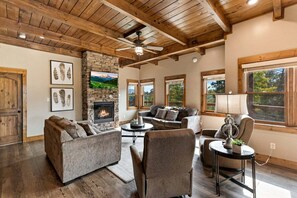 A spacious living room with a stone fireplace, wooden ceiling beams, and large windows offering views of the outdoors. - Moonlit Lodge - Hot Tub, Modern Vibes & Firepit (Sevierville)