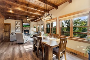 A spacious living area with a fireplace, wooden dining table, and large windows offering a view of the outdoors. - Moonlit Lodge - Hot Tub, Modern Vibes & Firepit (Sevierville)