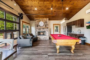 A room with a pool table, a fireplace, a stone wall, and a kitchen area. - Moonlit Lodge - Hot Tub, Modern Vibes & Firepit (Sevierville)