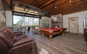 A spacious room with a pool table, leather chairs, a stone fireplace, and a large window overlooking a forest.