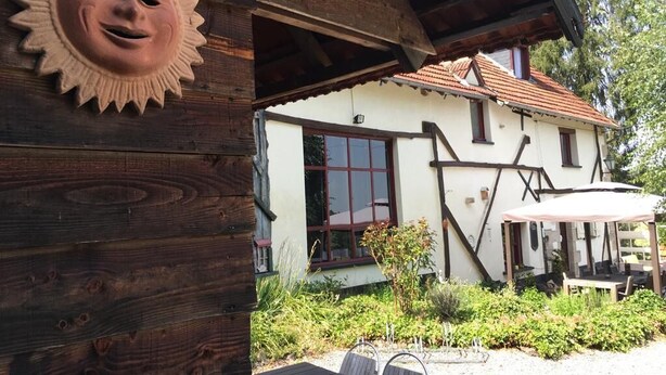 Een rustiek houten gebouw met een zonvormige decoratie, een overdekt terras en een raam met rode omlijsting.