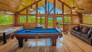 Un intérieur en bois avec une table de billard, un canapé et de grandes fenêtres donnant sur un paysage verdoyant.