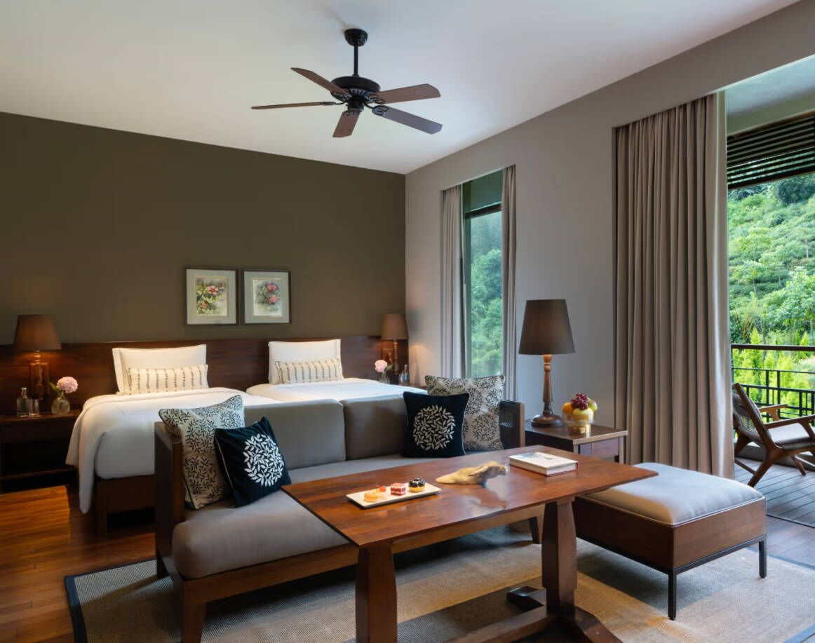 A hotel room with a bed, a sofa, a table with a lamp, and a view of greenery outside.
