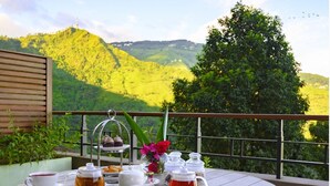 Une table ronde en bois, agrémentée d’un service à thé, d’un présentoir à gâteaux et d’un vase de fleurs, est placée sur un balcon qui offre une vue sur un paysage montagneux.